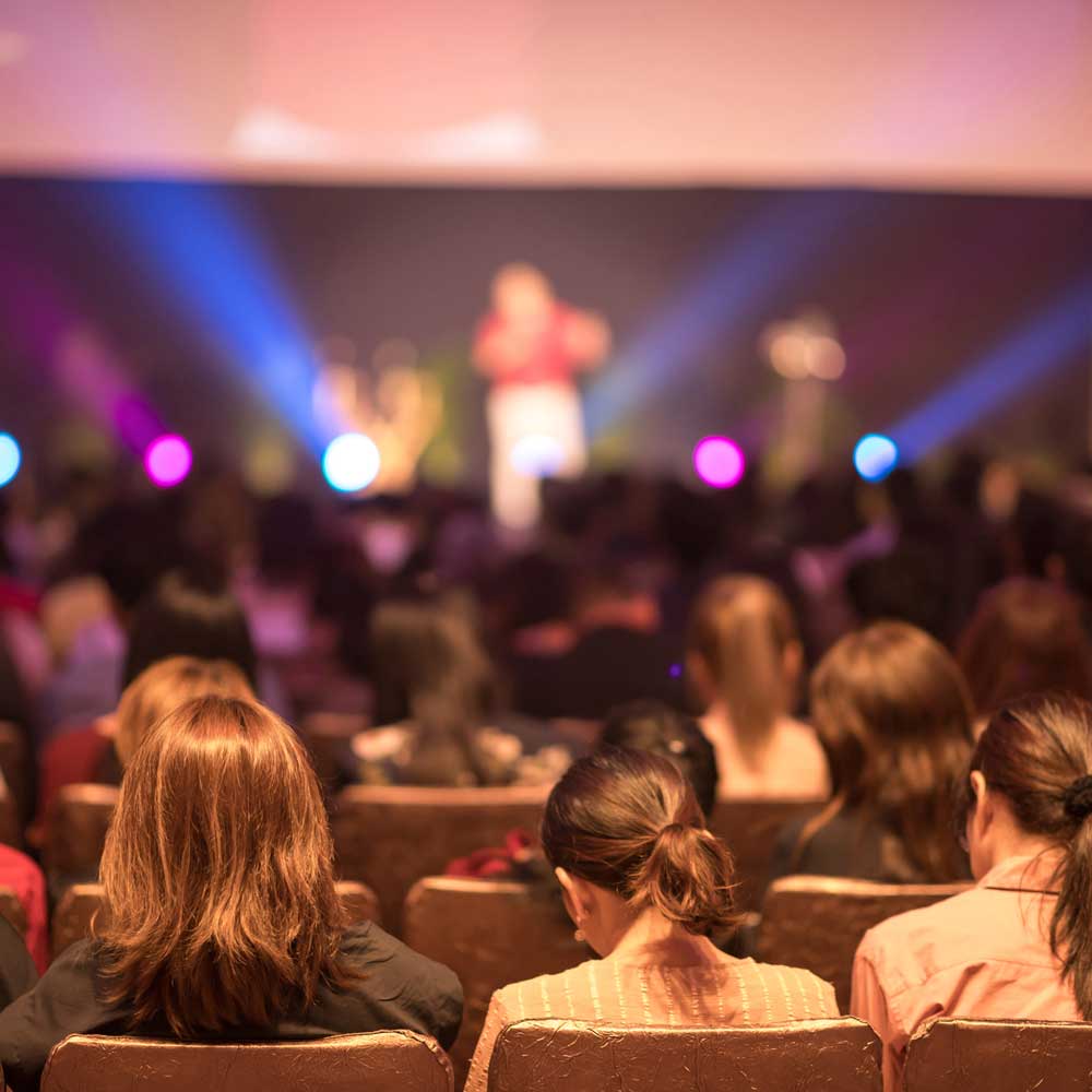 people seated in auditorium listening to speaker on stage